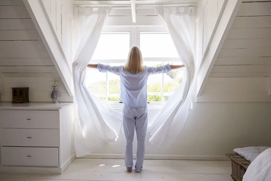 Eine Person in blauem Pyjama öffnet weiße Vorhänge vor einem Fenster in einem weißen Dachzimmer, dahinter grüne Bäume.