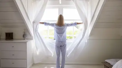Eine Person in blauem Pyjama öffnet weiße Vorhänge vor einem Fenster in einem weißen Dachzimmer, dahinter grüne Bäume.