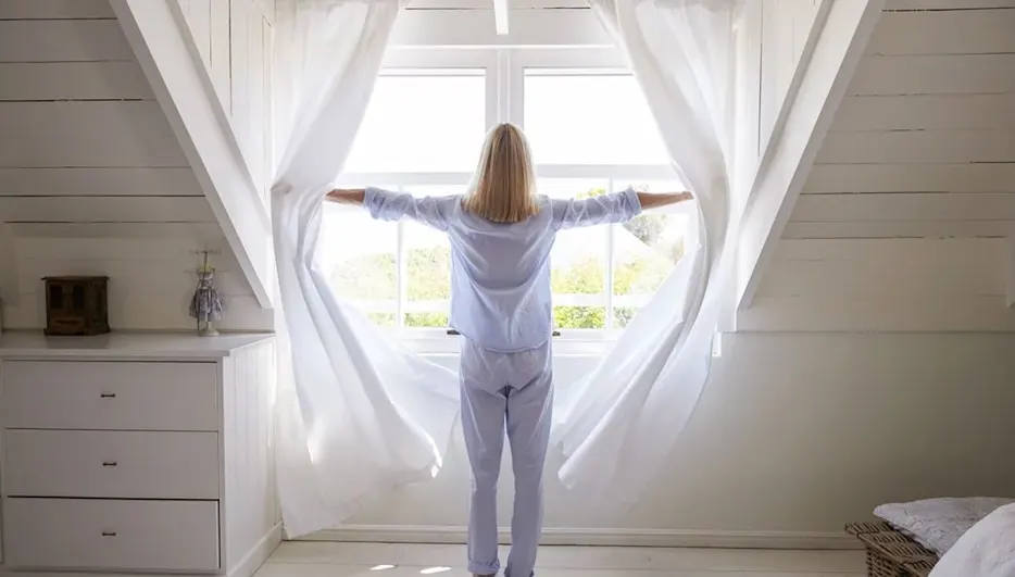 Eine Person in blauem Pyjama öffnet weiße Vorhänge vor einem Fenster in einem weißen Dachzimmer, dahinter grüne Bäume.