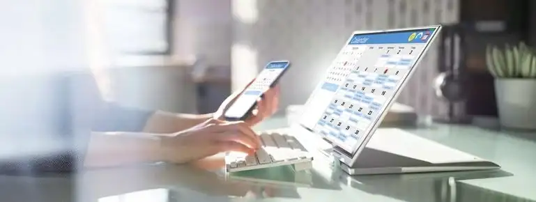Hände tippen auf weißer Tastatur, halten schwarzes Smartphone mit Kalender. Silberner Laptop zeigt digitale Kalenderansicht.