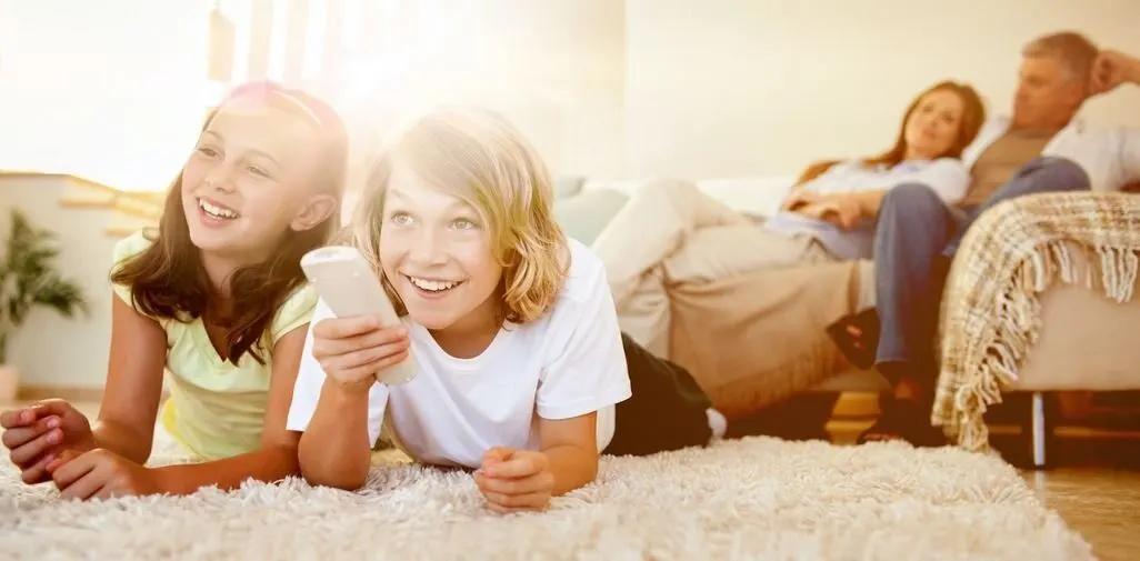 Zwei Kinder liegen auf dem Teppich und bedienen eine Fernbedienung, im Hintergrund die Eltern am Sofa
