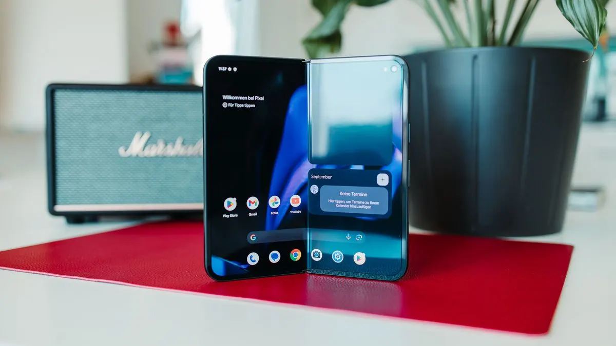 Offenes, dunkles Google Pixel 9 Pro Fold Smartphone auf roter Matte. Links App-Symbole, rechts Kalender-Widget. Im Hintergrund: Marshall-Lautsprecher, Pflanzentopf.