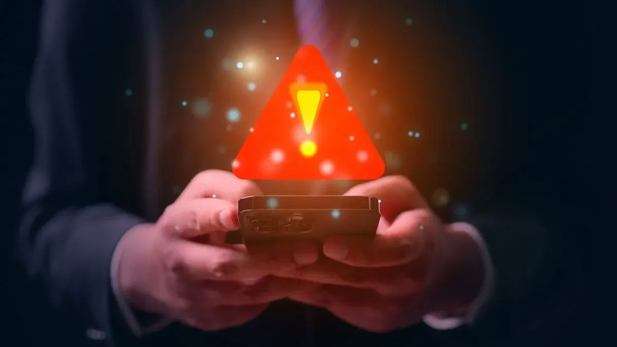 Hände halten ein dunkles Smartphone mit Daumen auf dem Bildschirm. Ein leuchtendes rotes Warndreieck mit gelbem Ausrufezeichen schwebt darüber.