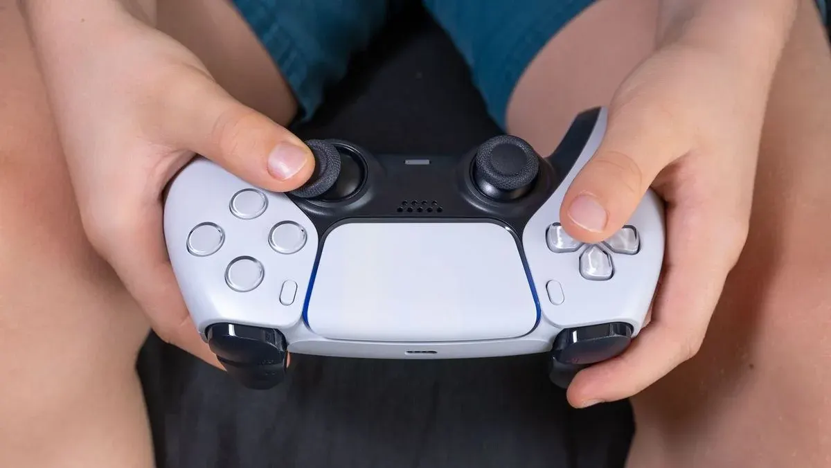Hände halten einen weiß-schwarzen Gamecontroller mit blauen Lichtstreifen. Daumen liegen auf den Joysticks.