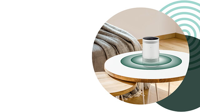 Ein runder Tisch mit einem zylindrischen Lautsprecher in einem Wohnzimmer. Wellenmuster im Hintergrund.