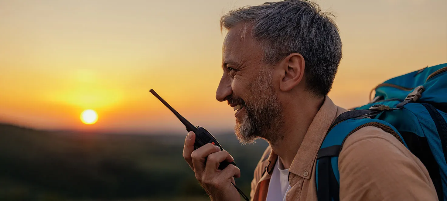 Ein Wanderer mit Rucksack hält ein Funkgerät vor einem Sonnenuntergang.