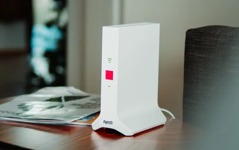 Weißer Fritz!Box Router auf Holztisch. Rote, grüne WLAN- und grüne Strich-Lichter leuchten. Zeitschriften daneben, unscharfe Möbel im Hintergrund.