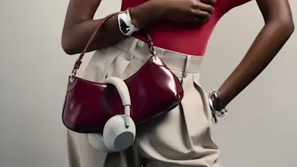 Glänzende weinrote Tasche, beigefarbene Kopfhörer, getragen von Person in rotem Oberteil, beiger Hose und Silberarmbändern.