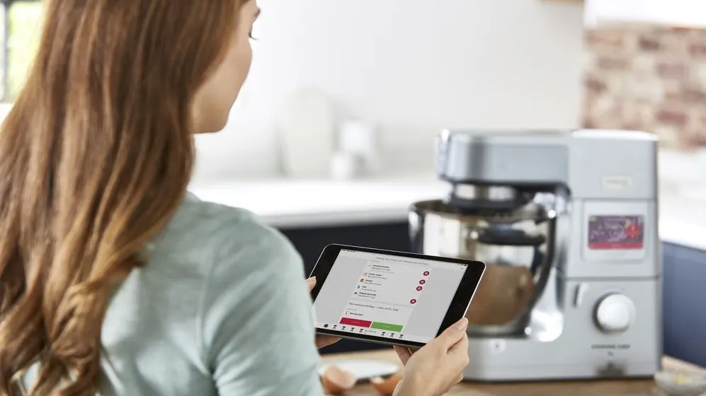 Person hält Tablet mit Rezept. Eine silberne Küchenmaschine mit Glasschüssel, Teig und Eiern steht auf einem Holztresen in heller Küche mit Ziegelwand.