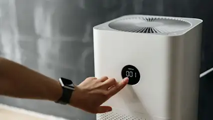 Zeigefinger einer Hand schaltet via Display einen Luftreiniger ein, der vor einer Glaswand steht.