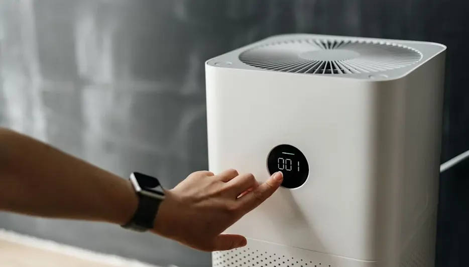 Zeigefinger einer Hand schaltet via Display einen Luftreiniger ein, der vor einer Glaswand steht.