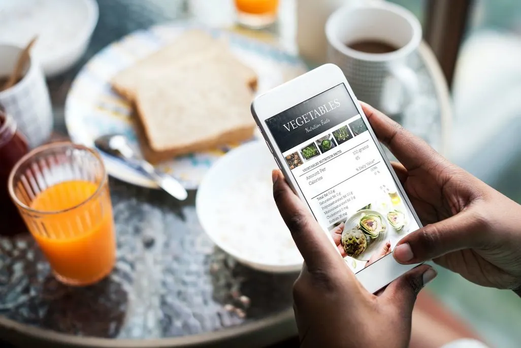 Eine Hand haelt ein Smartphone mit einer Essensapp am Fruehstueckstisch