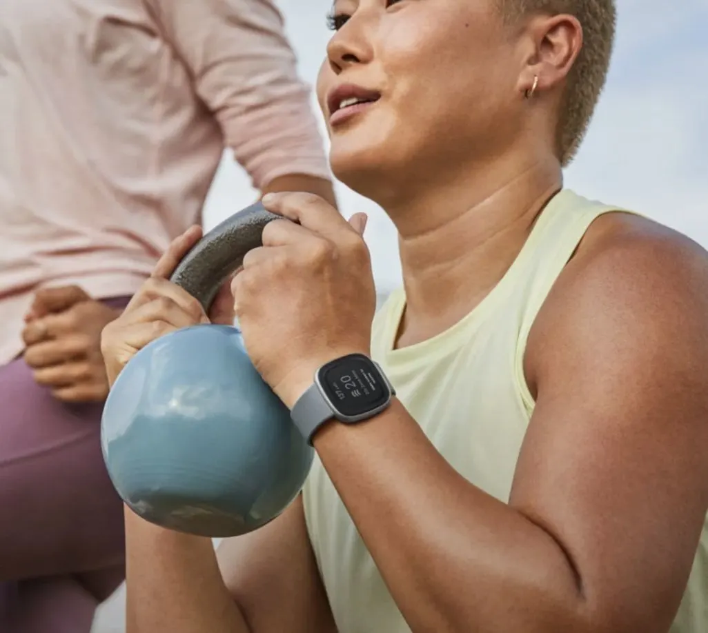 Person in Sportkleidung hebt eine blaue Kettlebell und trägt eine Fitbit Smartwatch.