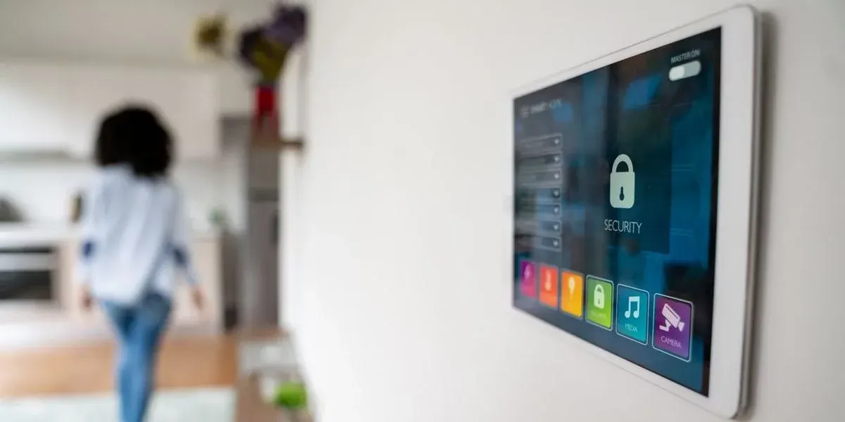 Ein Smart-Home-Tablet an einer weißen Wand zeigt "SECURITY" mit einem Schlosssymbol. Eine Person in weißem Hemd und blauen Jeans geht in eine unscharfe moderne Küche.