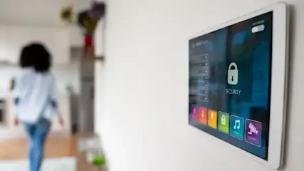 Ein Smart-Home-Tablet an einer weißen Wand zeigt "SECURITY" mit einem Schlosssymbol. Eine Person in weißem Hemd und blauen Jeans geht in eine unscharfe moderne Küche.