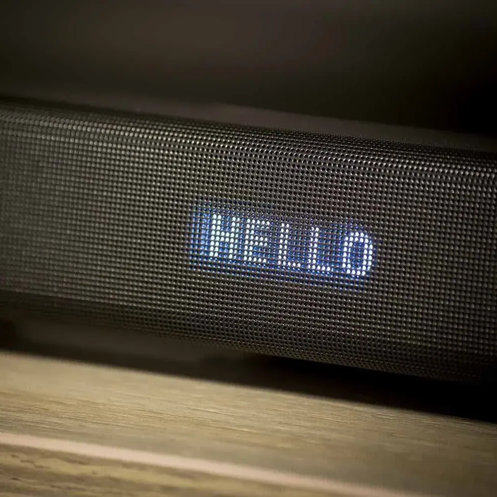 Schwarzer tragbarer Bluetooth-Lautsprecher mit grauem Textilgeflecht liegt auf Holzoberfläche. Blaue LED-Anzeige zeigt "HELLO"-Text. Moderne Elektronik.