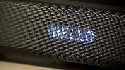 Schwarzer tragbarer Bluetooth-Lautsprecher mit grauem Textilgeflecht liegt auf Holzoberfläche. Blaue LED-Anzeige zeigt "HELLO"-Text. Moderne Elektronik.