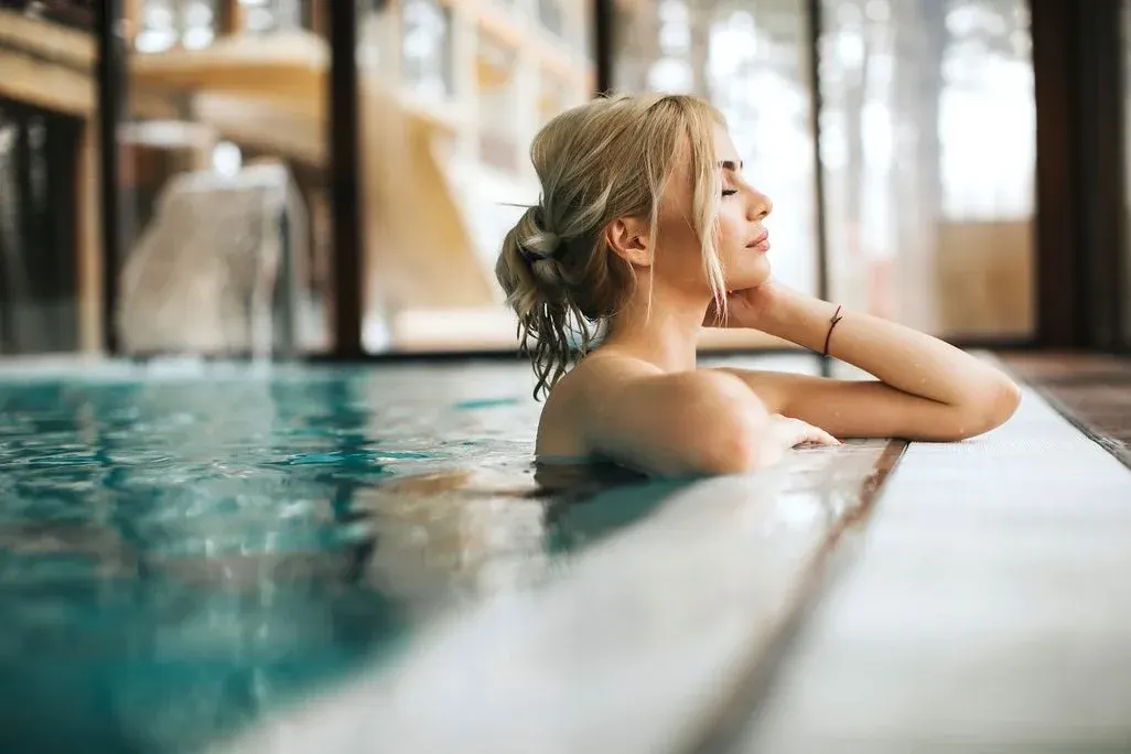 Eine Person entspannt sich in einem blauen Pool, die Arme auf dem weißen Beckenrand. Augen geschlossen, mit Fenstern im unscharfen Hintergrund.