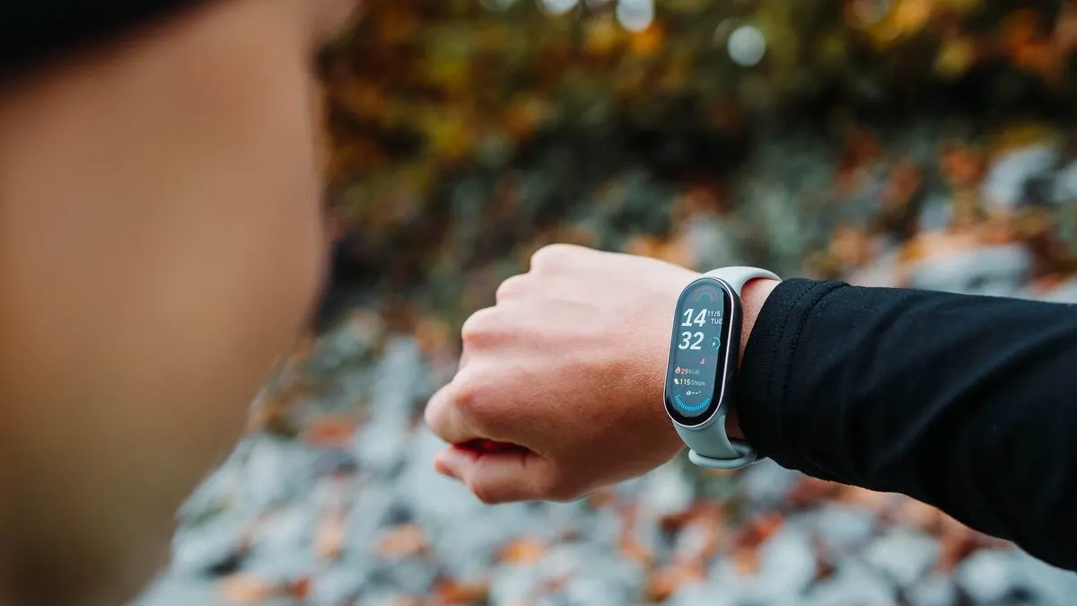 Eine Person prüft einen hellgrauen Fitness-Tracker am Handgelenk, der "14:32" und "115 Schritte" anzeigt. Im Hintergrund sind unscharfe Herbstblätter.