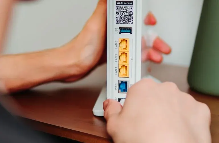 Weiße Router-Rückseite mit QR-Code, blauem USB-, gelben LAN- und blauem WAN-Port. Eine Hand interagiert, auf braunem Tisch, heller Wand, grüne Vase.