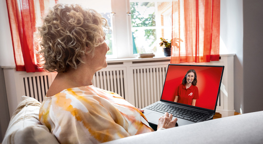 Frau sitzt auf Sofa mit Laptop. Auf dem Bildschirm ist eine Frau im roten Hemd zu sehen. Fenster und Vorhänge im Hintergrund.