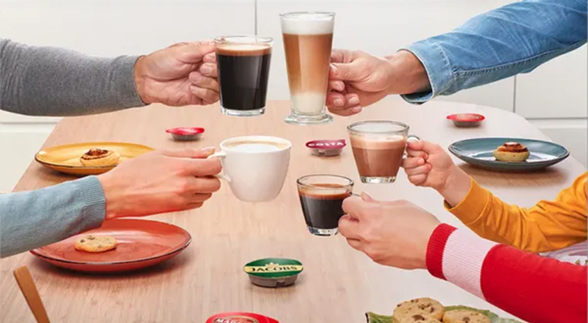 Auf einem Tisch stehen Kaffeetassen, Gläser und Teller mit Gebäck, die von mehreren Händen gehalten werden. Kapseln sind verstreut.