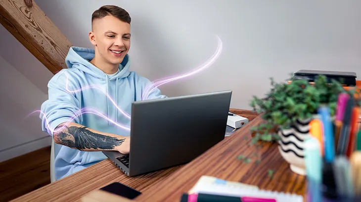 Junger Mann im hellblauen Hoodie arbeitet lächelnd an einem Laptop an einem Holztisch; leuchtende Linien symbolisieren digitale Vernetzung.