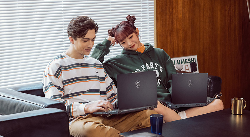 Zwei Personen sitzen mit Laptops auf einem Sofa vor einem Fenster mit Jalousien. Auf dem Tisch stehen eine Tasse und ein Glas.