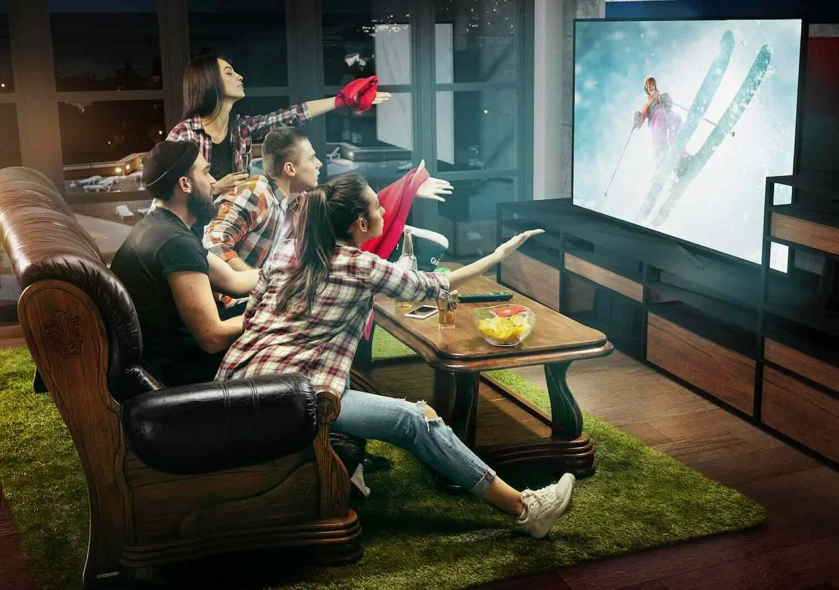  Gruppe junger Leute sitzt gespannt vor einem großen Fernseher im Wohnzimmer und feuert beim Anschauen eines Skirennens die Sportlerin auf dem Bildschirm an.