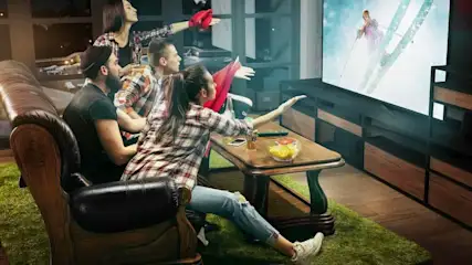 Gruppe junger Leute sitzt gespannt vor einem großen Fernseher im Wohnzimmer und feuert beim Anschauen eines Skirennens die Sportlerin auf dem Bildschirm an.