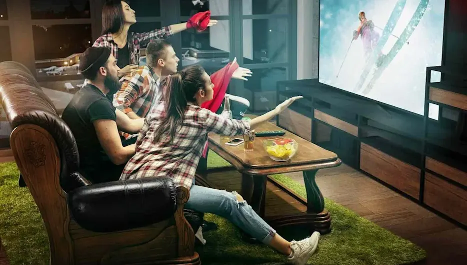 Gruppe junger Leute sitzt gespannt vor einem großen Fernseher im Wohnzimmer und feuert beim Anschauen eines Skirennens die Sportlerin auf dem Bildschirm an.