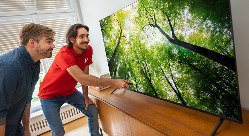 Zwei Männer schauen einen großen Fernseher, der auf einem Holzschrank steht. Auf dem Bildschirm ist ein grüner Wald zu sehen.