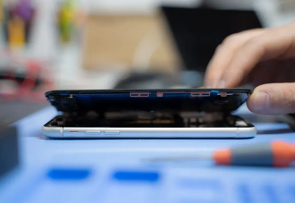 Hand öffnet silbernes Smartphone auf blauer Matte. Schwarzer Bildschirm ist angehoben, interne Teile sichtbar. Schraubendreher im Vordergrund.
