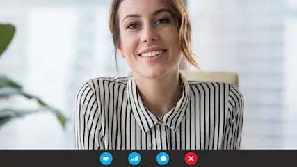 Frau in gestreifter Bluse sitzt bei einem Videotelefonat vor einem unscharfen Hintergrund. Unten im Bild sind die Symbole einer Videochat-Software sichtbar.