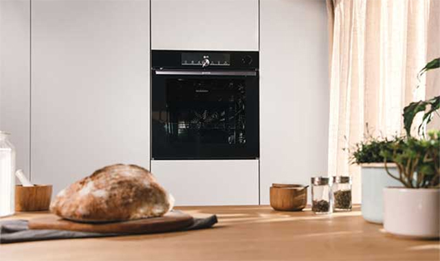 Ein Brot auf einem Holzbrett, ein Backofen in der Wand, Gewürze und Pflanzen auf einer Arbeitsfläche.