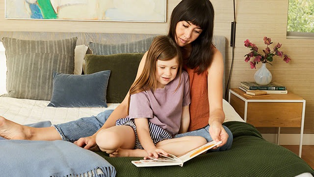 Eine Frau und ein Kind sitzen auf einem Bett und lesen ein Buch. Kissen und eine Nachttischlampe sind auch zu sehen.