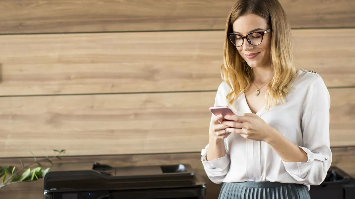 Frau steht vor einem Drucker und bedient ein Smartphone.