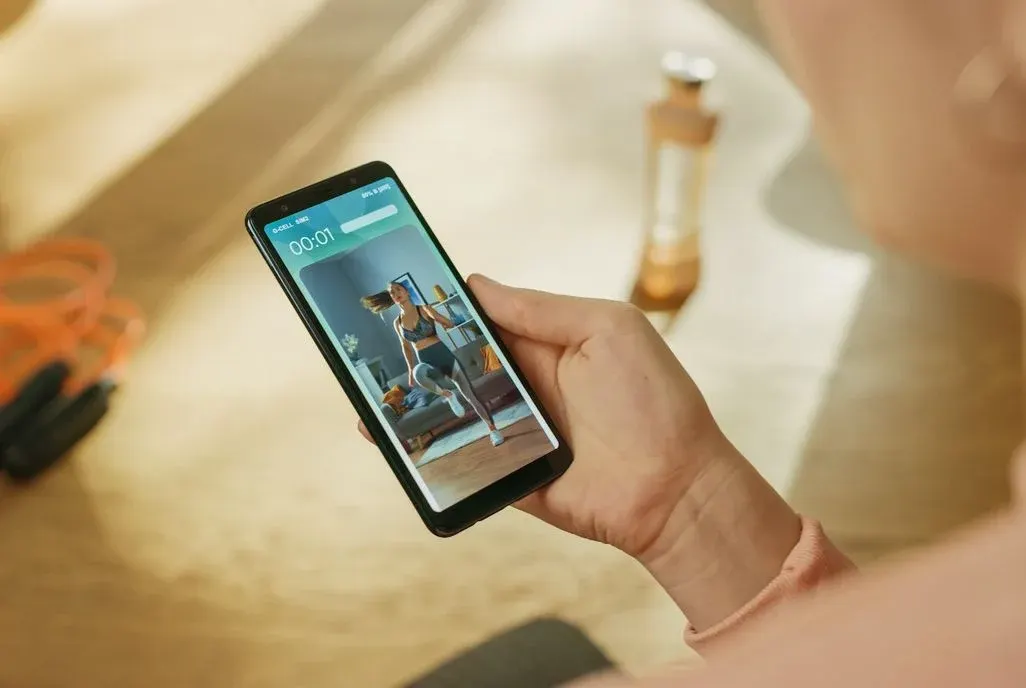 Hand hält Smartphone mit Fitness-Video: Eine Person in grauer Sportkleidung macht Übungen. Im Hintergrund sind ein Springseil und eine Flasche.
