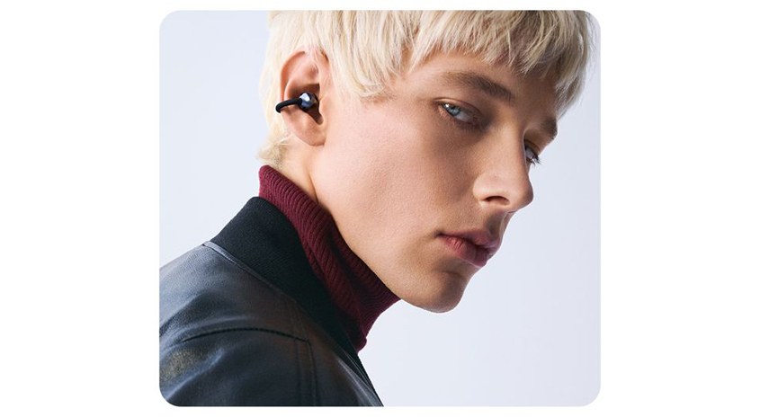 Nahaufnahme einer Person mit kurzem, blondem Haar, die schwarze In‑Ear-Kopfhörer trägt und seitlich in die Kamera blickt, vor hellem Hintergrund.