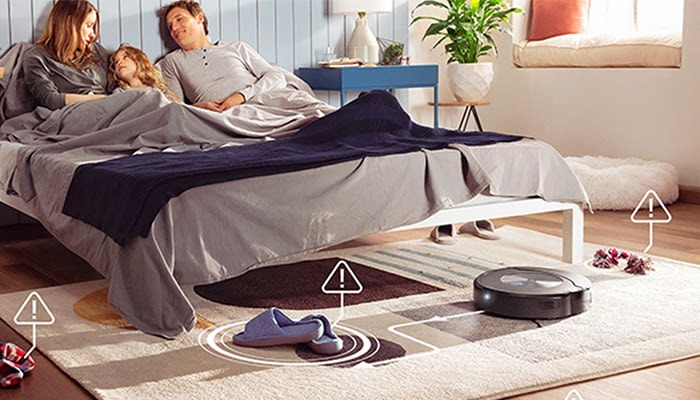 Familie liegt im Bett, während ein Roboterstaubsauger auf einem Teppich arbeitet. Neben dem Bett stehen Tisch und Pflanze.