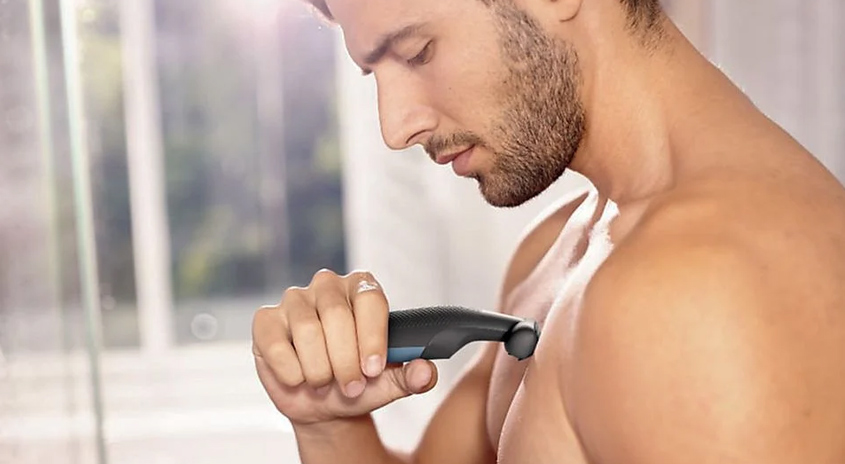 Ein Mann trimmt seine Brusthaare mit einem schwarzen und blauen Trimmer vor einem Fenster.