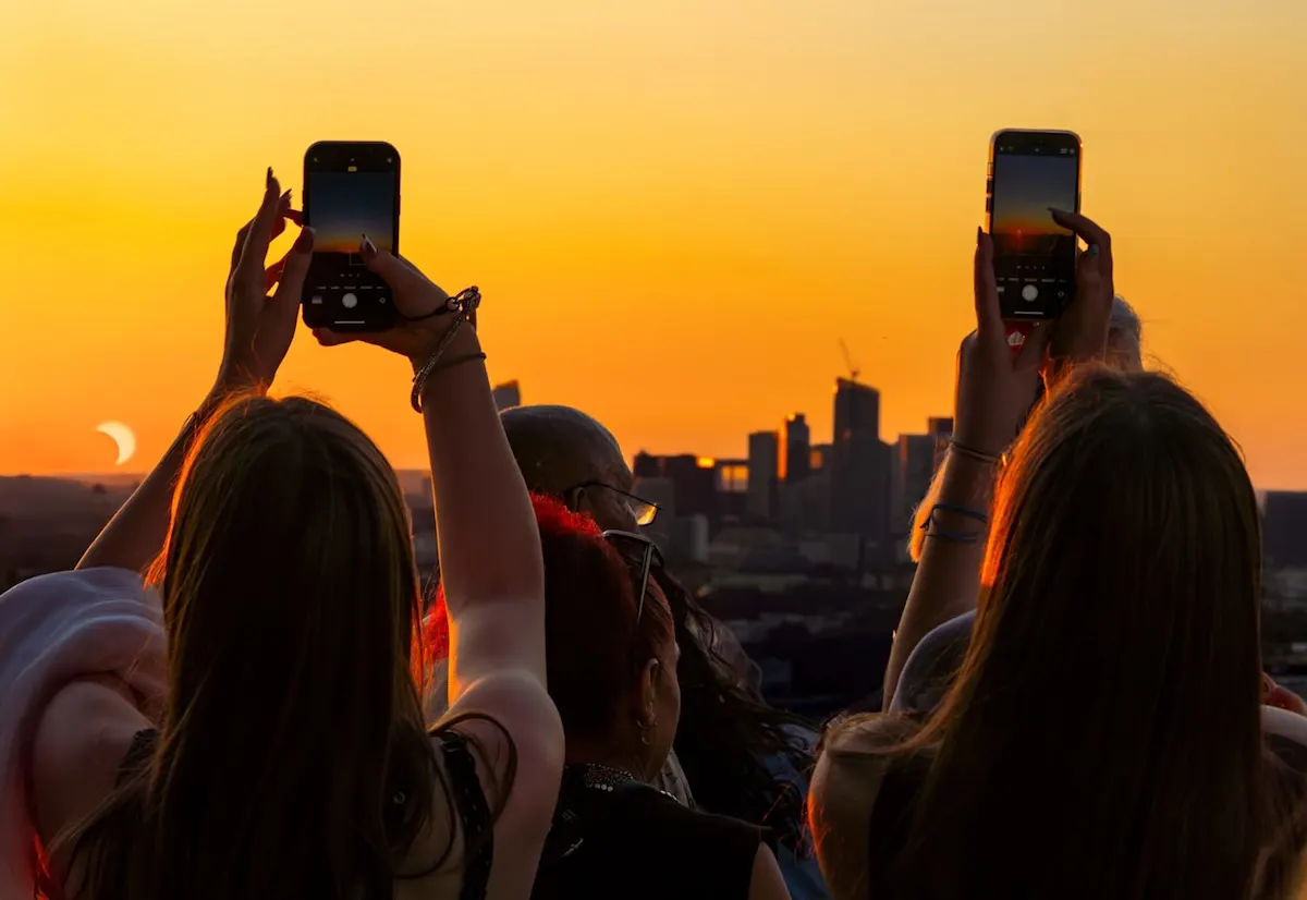 Mehrere Personen fotografieren vor orangem Himmel mit dem Smartphone den im Bildhintergrund zu sehenden Sonnenuntergang.