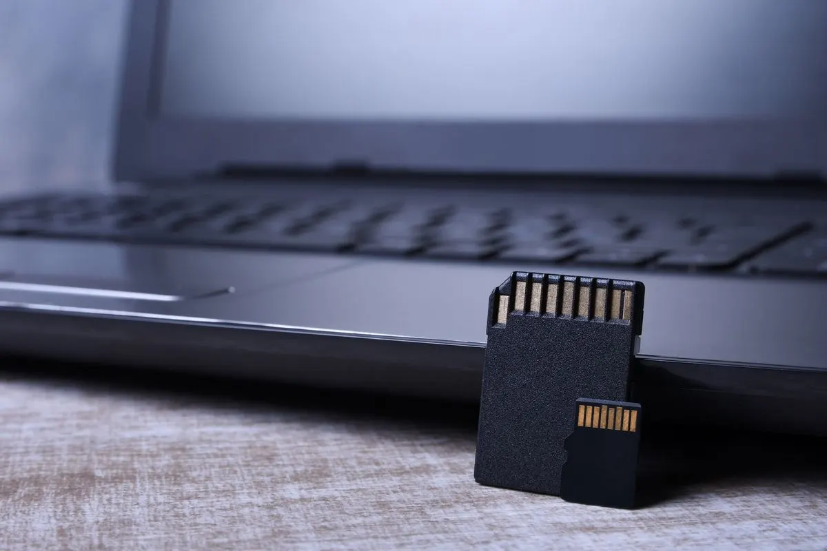 Zwei schwarze Speicherkarten mit goldfarbenen Kontakten stehen auf hellem Holz vor einem unscharfen grauen Laptop.
