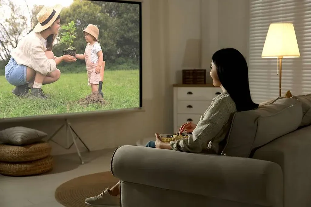 Frau sitzt abends entspannt auf dem Sofa und schaut eine Beamer-Projektion, auf der eine Mutter mit Kind gemeinsam draußen einen jungen Baum pflanzt.