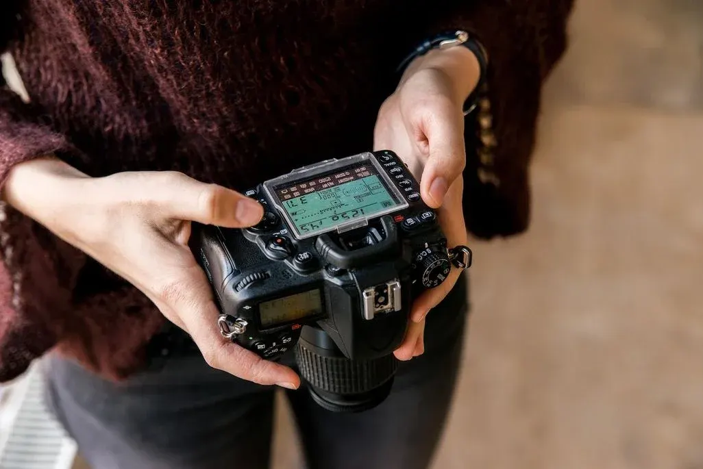 Person in dunkelrotem Pullover hält schwarze DSLR-Kamera, passt das grüne Display an. Schwarze Hose, heller, unscharfer Hintergrund.