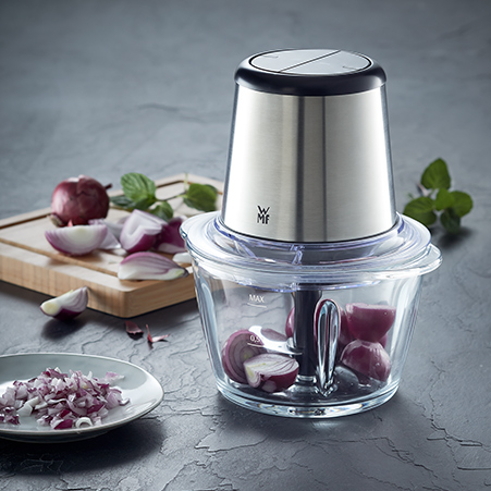 Silberner Zerkleinerer mit Glasschüssel, gefüllt mit roten Zwiebeln. Zwiebeln auf Brett und Teller im Hintergrund.