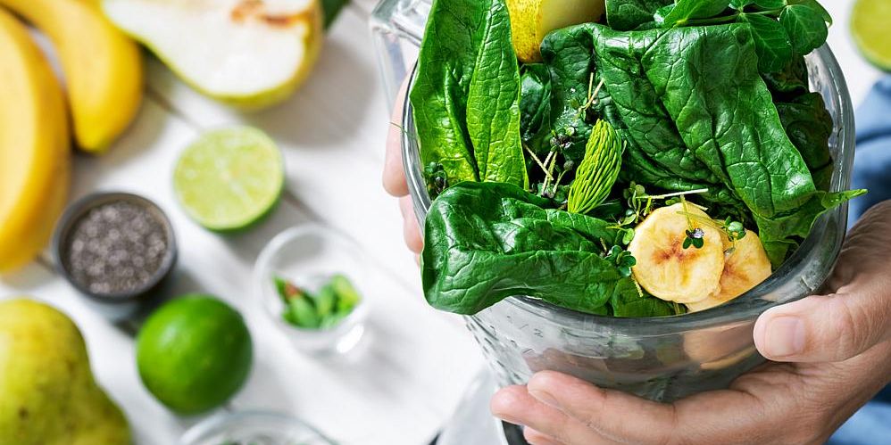 Hände halten einen Mixer mit frischem Spinat, Kräutern und Bananenscheiben, daneben liegen verschiedene Obstsorten.