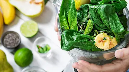 Hände halten einen Mixer mit frischem Spinat, Kräutern und Bananenscheiben, daneben liegen verschiedene Obstsorten.
