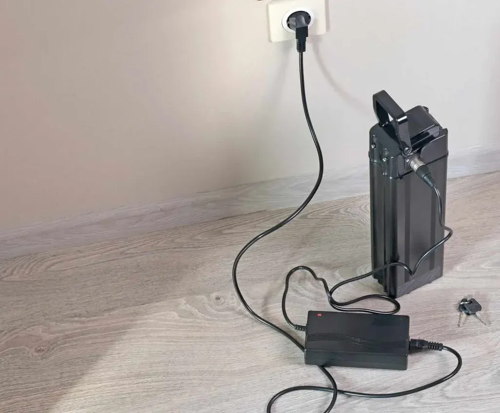 E‑Bike‑Akku steht auf dem Boden und ist über ein Ladegerät an eine Steckdose angeschlossen, daneben liegt ein Schlüssel.