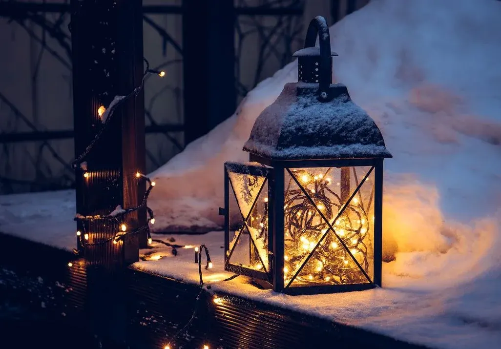 Person in weißem Pullover und grauen Leggings hält weiße Kaffeetasse. Rote Socken und funkelnde Lichterketten umgeben sie.